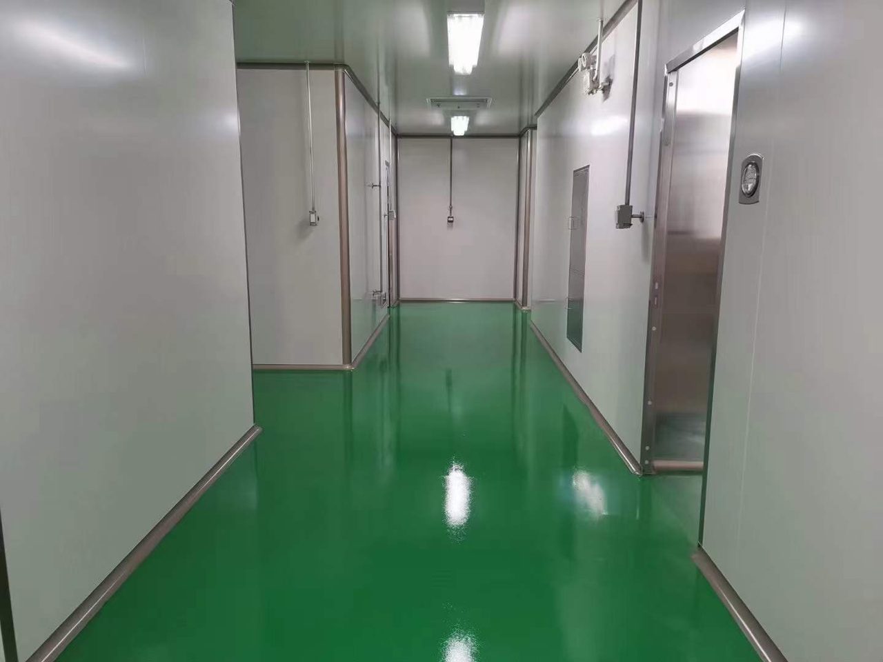 厂房车间环氧地坪施工实拍——环氧自流平,厂房车间内的绿色环氧自流平通道,两侧为车间墙面与金属门,顶部有照明灯具
